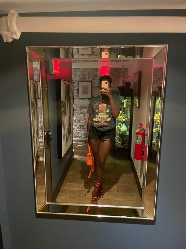 A woman taking a mirror selfie in a hallway with brick walls and artwork, wearing a t-shirt, shorts, and holding a bag.