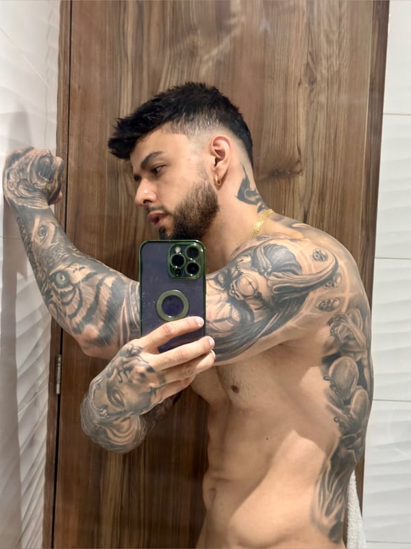 A muscular tattooed man flexing his arm in a bathroom mirror selfie, shirtless and showing off his physique.