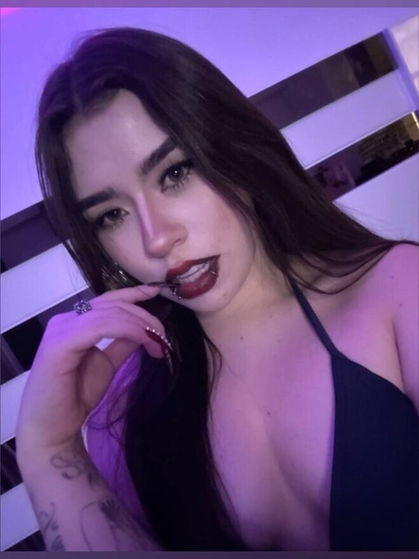 A woman with long dark hair and bold makeup posing close-up in purple lighting against a reflective surface.