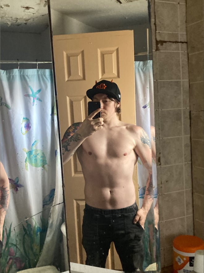 A young man with tattoos standing shirtless in a bathroom mirror selfie, wearing a black cap and black pants.