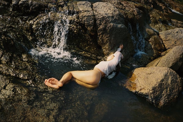 A young woman reclining on rocks near a small waterfall in a natural outdoor setting, wearing a white shirt.