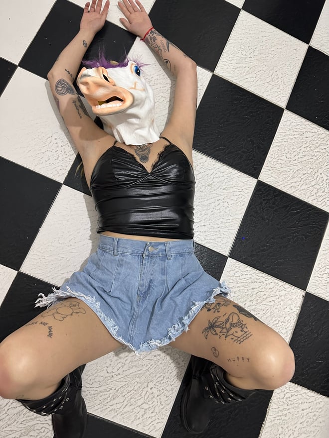 A person lying on a checkered floor wearing a unicorn mask, black top, shorts, and boots, with tattoos on arms and legs.