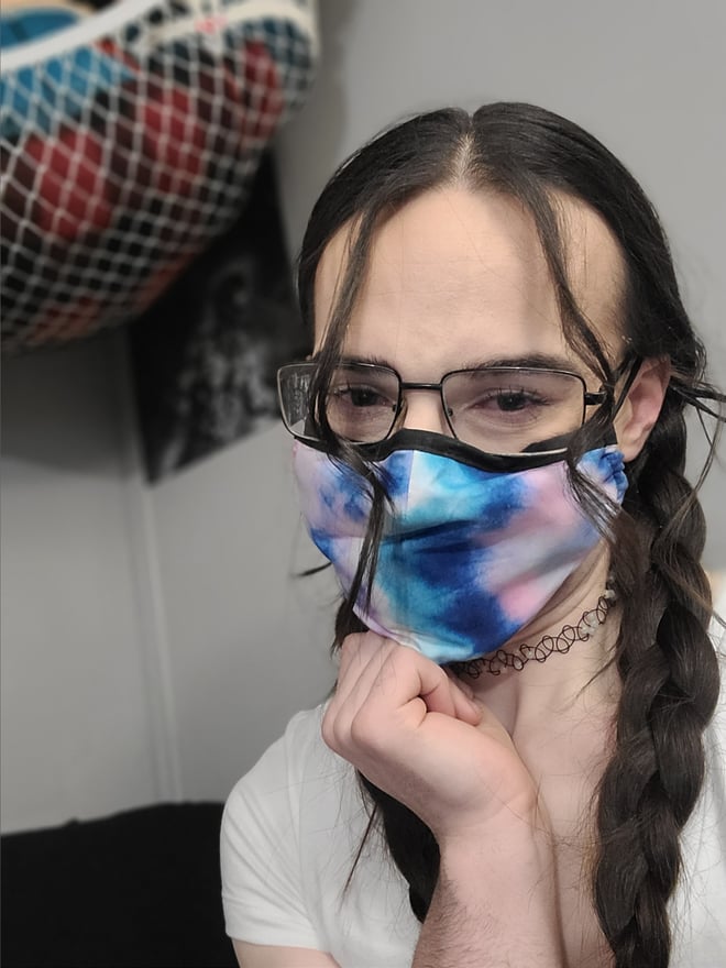 A person with glasses and a colorful face mask, wearing a choker and white top, posing with a hand on chin against a dark background with a netted wall hanging.