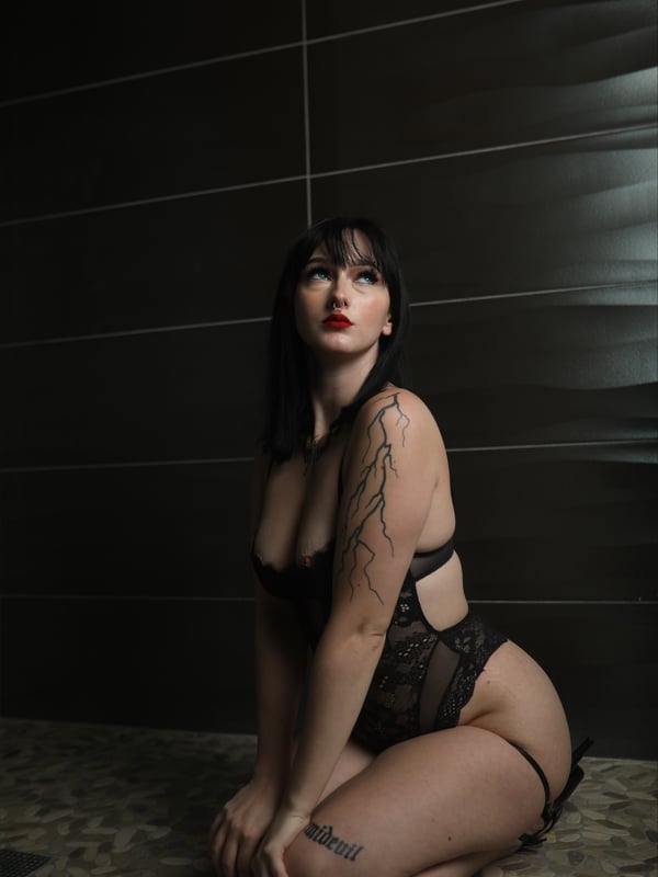 A woman with long dark hair kneeling on the floor against a dark tiled wall, dressed in a black lace top.