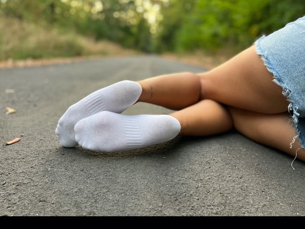 A person lying on a paved path in a green outdoor setting, wearing white socks, black shorts, and a blue denim skirt.