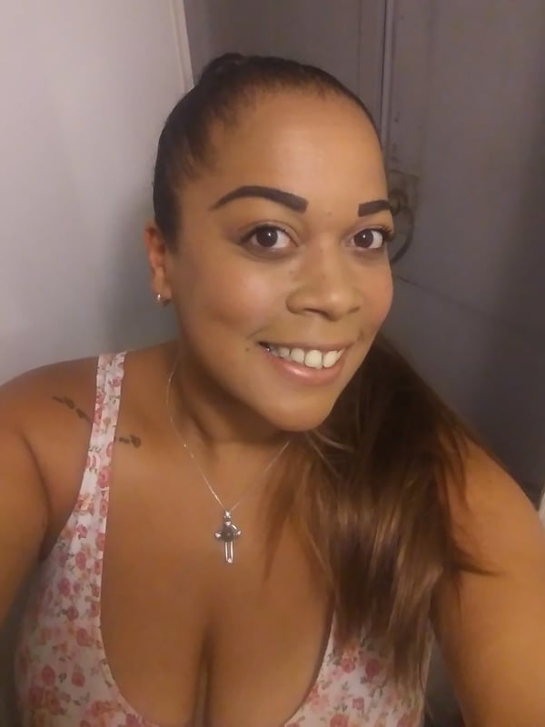 A smiling woman with a ponytail and dark hair, wearing a pink tank top and necklace against a plain background.