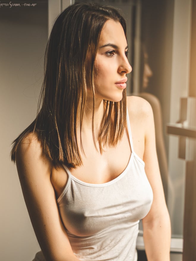 A woman with long brown hair wearing a white tank top stands by a window in a sunlit room.