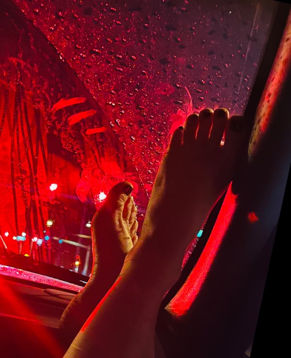 Close-up view of bare feet pressed against a car window with red neon lights and raindrops in the background at night.
