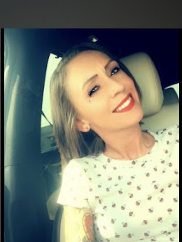 Young woman with blonde hair and tattoos on her arm smiling in a car, wearing a white polka-dot top.