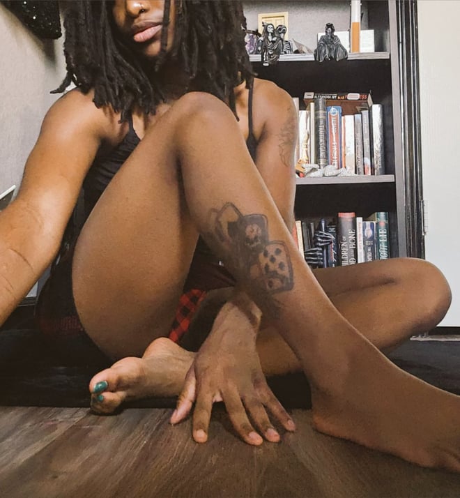 A woman with dreadlocks sits on the floor in a dimly lit room with bookshelves, posing with legs extended and feet visible.