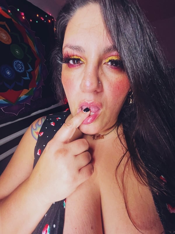 Close-up of a woman with dark hair and bold makeup pointing at her mouth, wearing a low-cut top with tattoos on her shoulder.