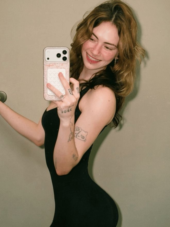 A young woman with wavy auburn hair poses outdoors at dusk in a patterned top, with visible tattoos and facial piercings.