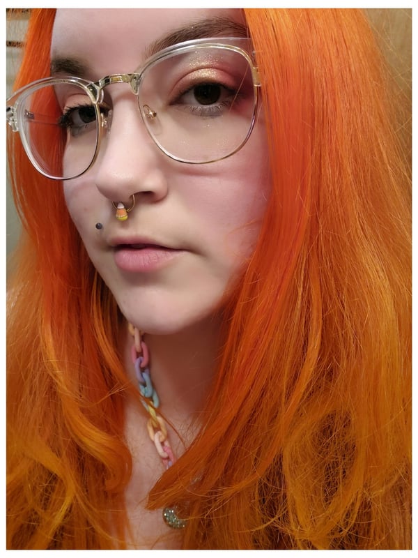 Woman with bright orange dyed hair, glasses, facial piercings, and a colorful chain necklace, posing with a subtle expression.