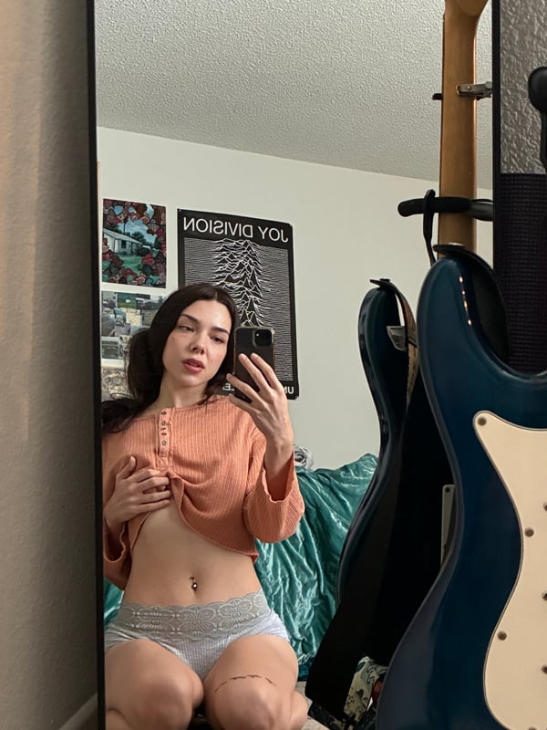 Young woman with long wavy dark hair smiling at the camera, wearing a gray long-sleeve crop top, standing in a colorful bedroom with posters on the walls.