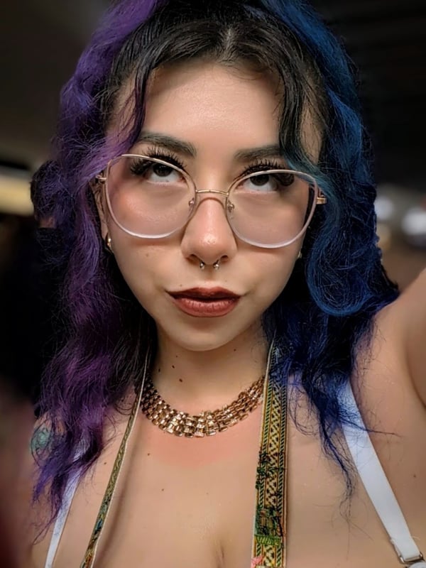 Young woman with dyed purple and black wavy hair, wearing clear frame glasses and a gold chain necklace, posing with a subtle smile in low light.