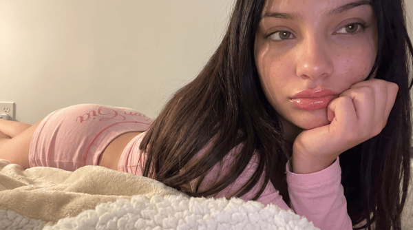 Young woman with long dark hair lying on a bed in a pink outfit, posing thoughtfully with her hand on her chin against a simple indoor background.
