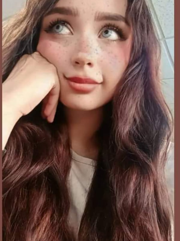 A young woman with long brown hair and freckles smiling softly while resting her chin on her hand.