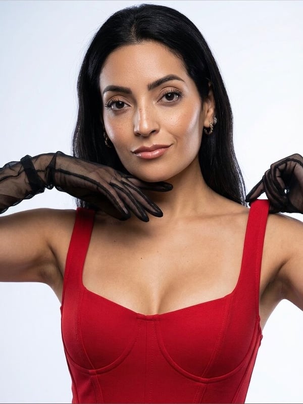 A woman with long dark hair posing confidently in a red sleeveless top and black lace gloves against a plain background.