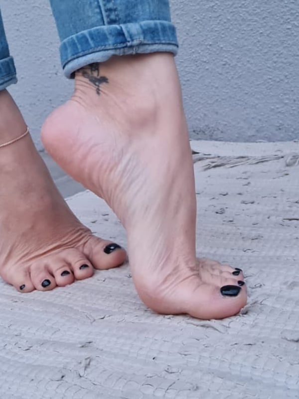 Close-up of bare feet with dark painted toenails standing on a textured surface, wearing slim jeans.