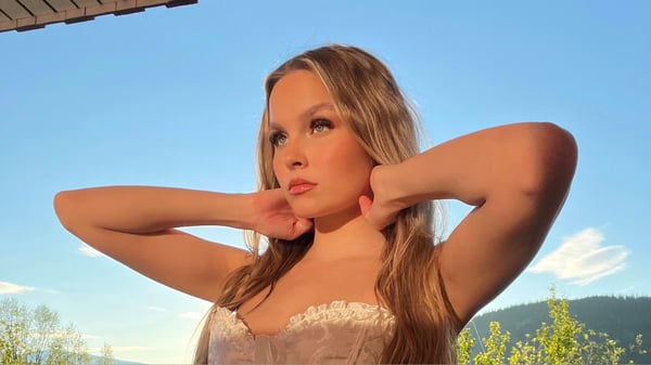 Young woman with long blonde hair posing confidently outdoors with arms raised, wearing a light-colored lace top against a scenic mountain backdrop.