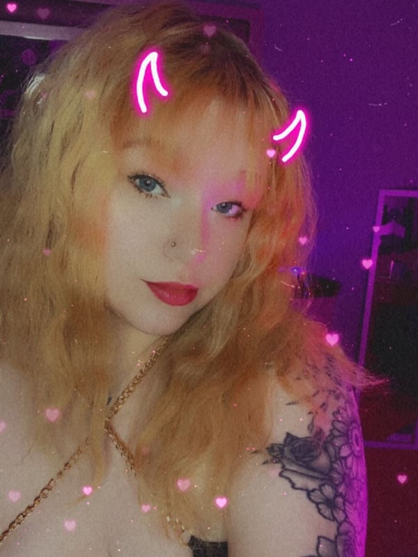Young woman with blonde hair, devil horns filter, and tattoos posing in a dimly lit purple room.