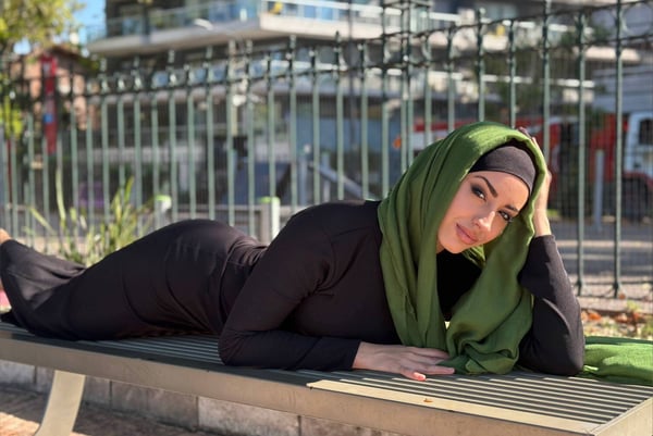 A woman wearing a green hijab and black dress reclines on a bench in an outdoor urban setting with fences and buildings in the background.