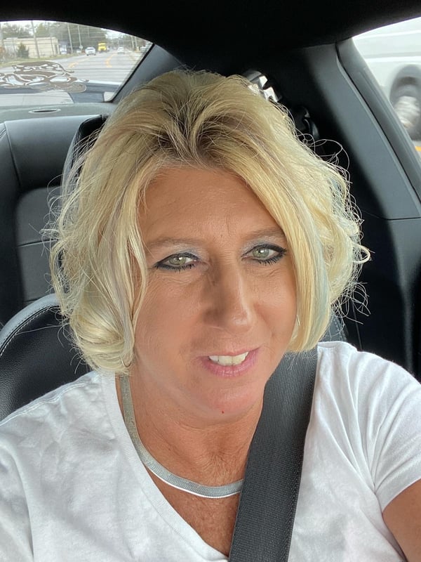A smiling woman with blonde hair sitting in a car, wearing a white top.
