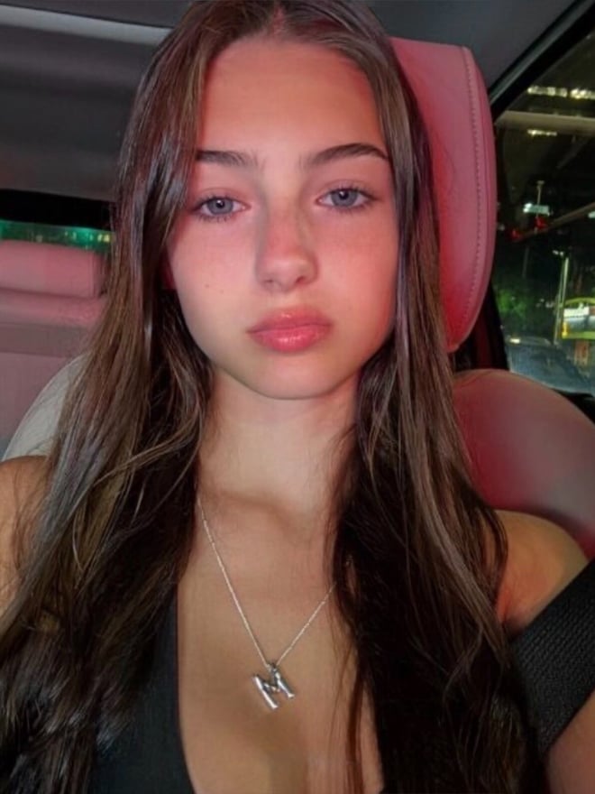 A young woman with long dark hair posing for a car selfie, wearing a dark top and necklace.