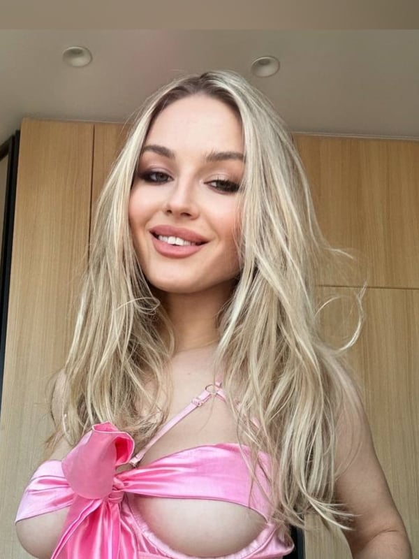 A woman with long blonde hair smiling at the camera, wearing a pink ribbon top, standing indoors near wooden doors.