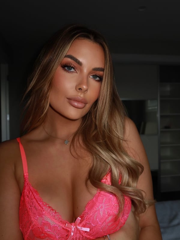 A woman with long blonde hair posing confidently in a pink lace top indoors with soft lighting.