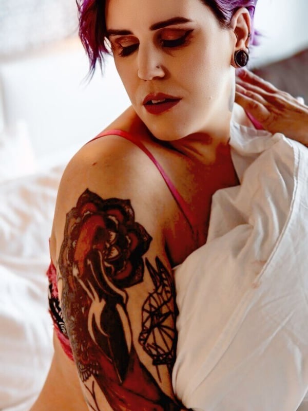 Close-up portrait of a woman with short purple hair, piercings, and tattoos, posing thoughtfully on a bed while holding a white sheet.
