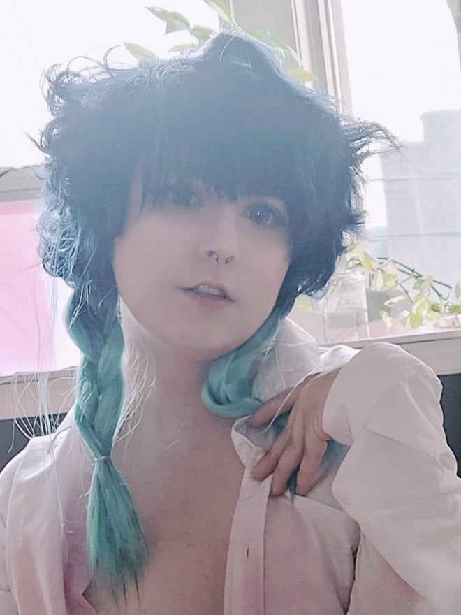 Person with teal braided hair and nose piercing, wearing an open white shirt, posing near a window with plants and city view in the background.