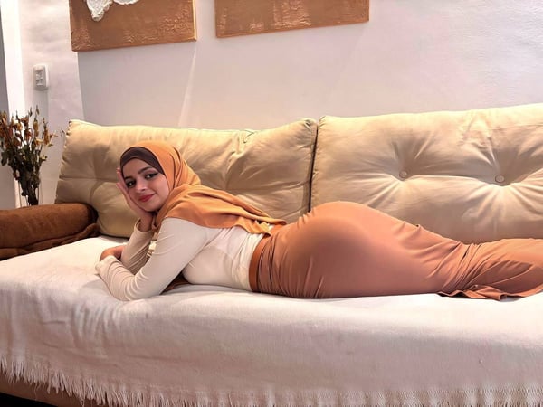 A woman in a hijab and long modest clothing reclines gracefully on a couch in a cozy room with artistic wall decor and dried flowers.