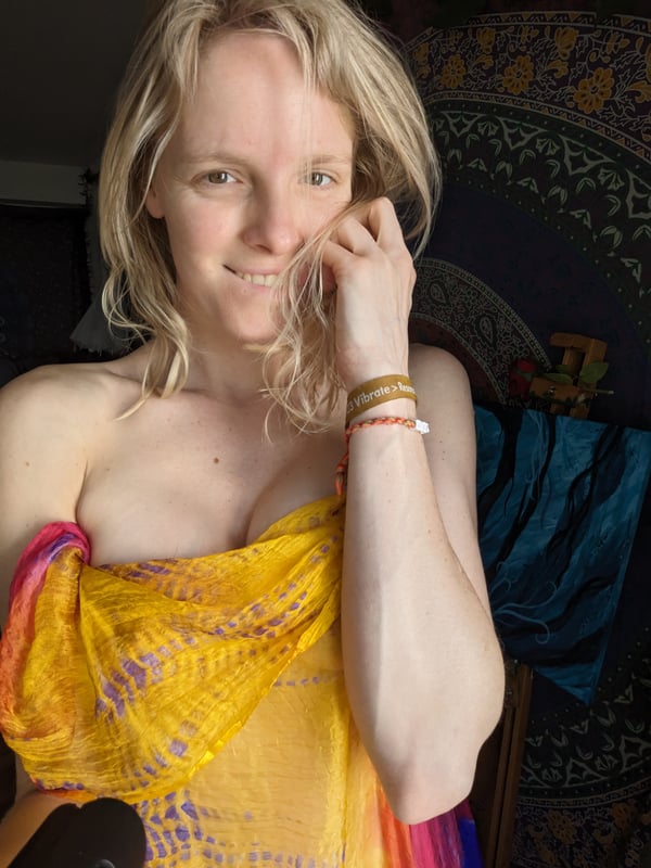 Blonde woman smiling with hand on face, draped in a colorful yellow and purple scarf, against a tapestry backdrop.
