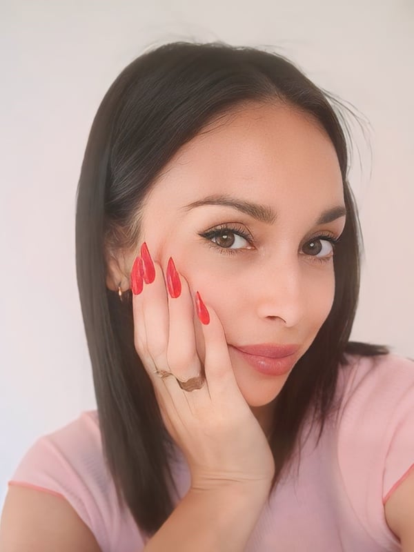 A woman with long dark hair and red nails posing with her hand on her cheek, wearing a pink top.