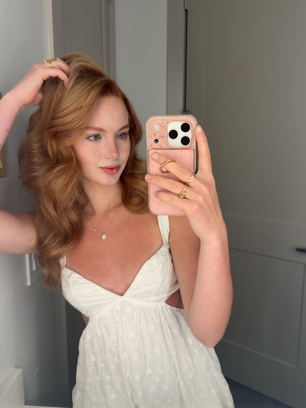 Young woman with blonde hair taking a selfie in a black halter top inside a bedroom with warm lighting.