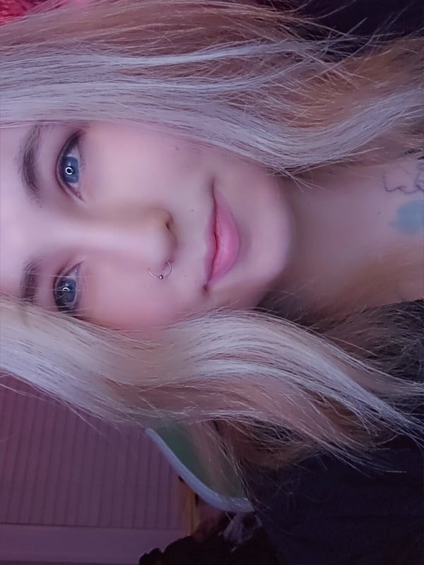 Close-up selfie of a young woman with long blonde hair, blue eyes, and subtle makeup, smiling softly in a dimly lit pink-toned room.