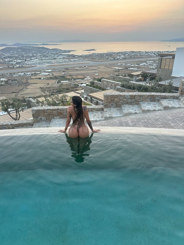 A woman sitting by a pool edge at sunset, overlooking a scenic coastal landscape with sea, hills, and distant buildings.