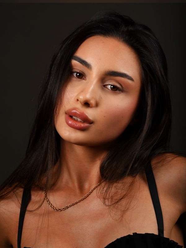 A close-up portrait of a woman with long dark hair wearing a black top and gold necklace.