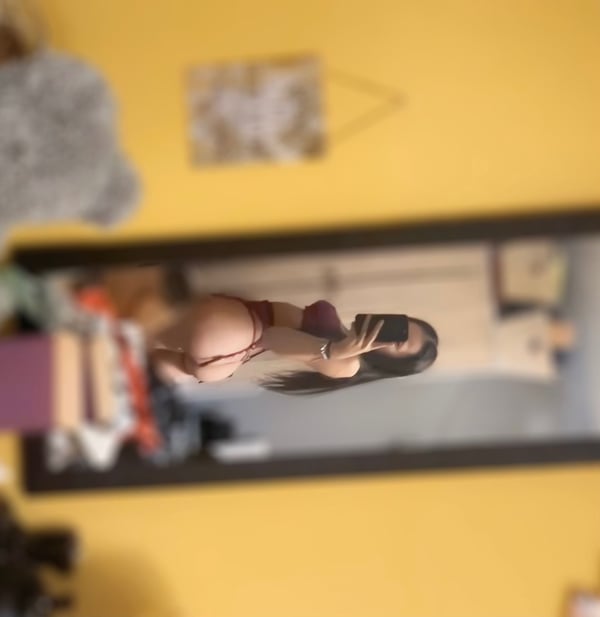 A young woman with long dark hair stands in front of a mirror in a yellow-walled room, wearing lingerie and holding a phone.
