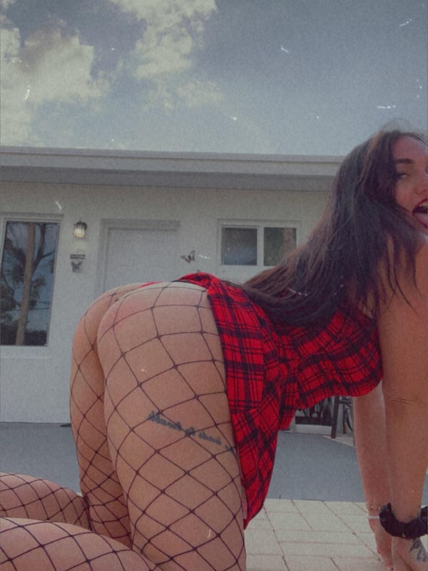 A young woman in a plaid dress and fishnet stockings posing playfully outdoors in front of a building under a cloudy sky.