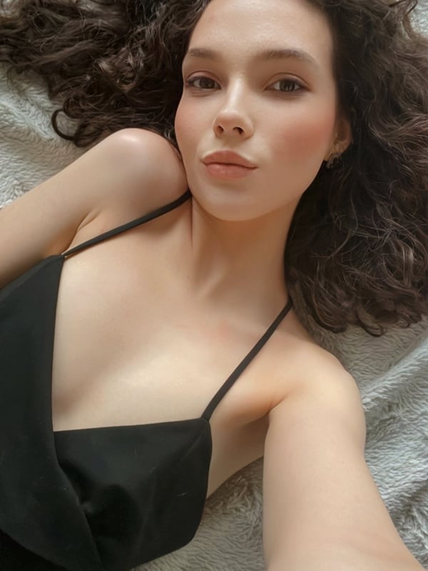 Young woman with long curly hair lying on a bed, wearing a black spaghetti strap top, with a subtle smile and natural makeup.
