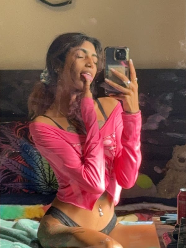 A young woman taking a selfie in a pink off-shoulder top and shorts, sitting on a colorful bed with tattoos visible.