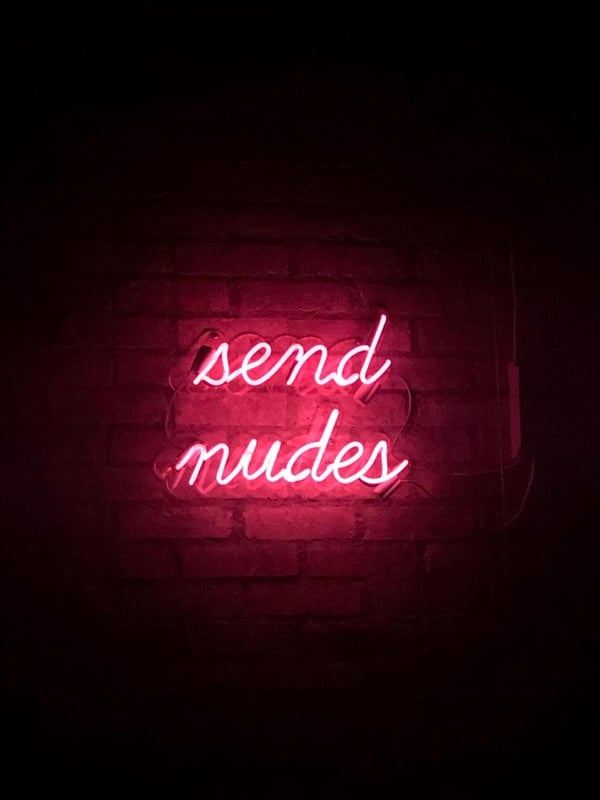 Neon sign with the text 'send nudes' illuminated in pink against a dark brick wall background.