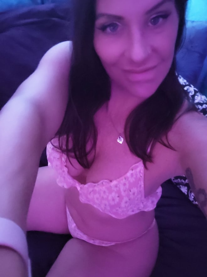 A woman with long dark hair posing on a bed in a pink lace top and bottoms, smiling at the camera in a cozy bedroom setting.