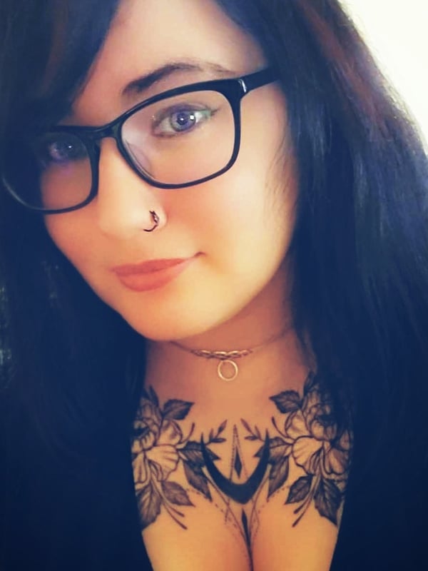 Close-up portrait of a woman with dark hair, glasses, and floral tattoos on her chest, smiling softly.