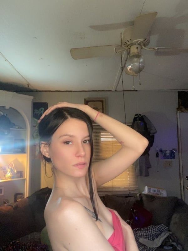 A young woman with long dark hair poses in a casual indoor setting with a ceiling fan and furniture in the background.