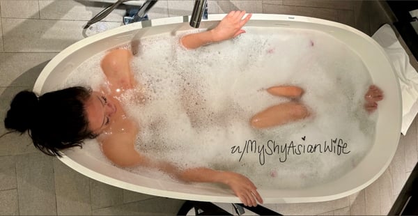 A woman relaxing in a bathtub filled with bubbles, with her hair tied up and arms resting on the edges.