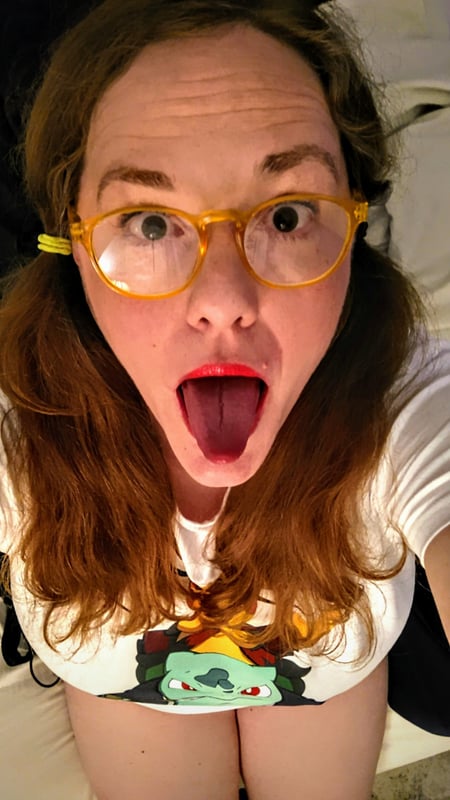 A woman with glasses and pigtails wearing a white top with a green character print, making a playful face with her tongue out in a close-up selfie.