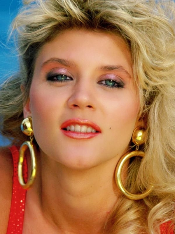 Close-up portrait of a woman with big blonde hair, bold makeup, large gold hoop earrings, smiling against a blue background.